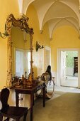 A yellow-painted hallway with a vaulted ceiling and a Baroque mirror above a wall table and a view through an open door