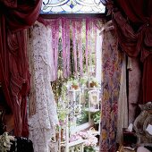 Dresses hanging on the wall in front of a opening with a fringe curtain and gathered red material with a view onto a conservatory