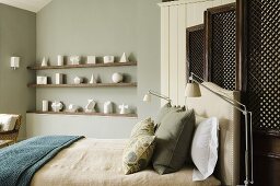 A double bed in front of a wooden paravent and geometric figures on a shelf in front of a grey wall