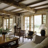 An elegant country house with a rustic wood beam ceiling and leather chairs in front of open swing doors leading to a terrace