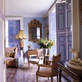 Baroque chairs and an occasional table in front of floor-to-ceiling window shutters and a mirror with a gold frame in a lilac-painted living room