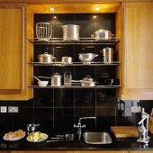 Kitchen cupboards with wooden doors with stainless steel pans on a wire shelf in a black-tiled niche