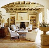 Blick durch Rundbogen in Mediterraner Wohnraum mit rustikaler Holzdecke und weiße Sesseln vor Kamin in Natursteinwand
