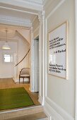 A white wood panelled wall in the hallway of a period building with a flight of stairs and a green bamboo mat on the wooden floor