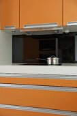 Küchenschrankausschnitt mit orangefarbenen Fronten und Edelstahltopf vor schwarzer Rückwand