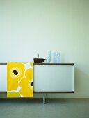 A detail of a modern, sitting room, retro styled sideboard cabinet, bright yellow and black cloth, glassware,