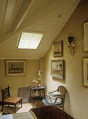 A detail of a country style bedroom, wood panelled sloping ceiling, bed, chairs, side table, skylight,