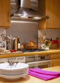 A detail of a modern kitchen with wood units, stainless steel extractor fan, hob and splash back, utensils