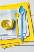 An arrangement of blue and yellow - a sticky tape dispenser and plastic spoons on a pile of tea towels