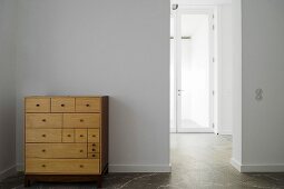 A simple wooden chest of drawers against a white wall and an open doorway with a view of a glass door