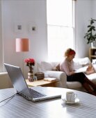 A laptop and a cup of coffee on a table with a woman in the background sitting on a sofa