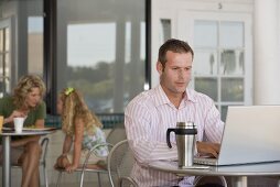 Man with laptop at bistro table, woman & child in background