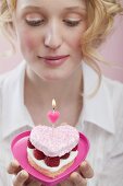 Young woman holding raspberry cake with candle in heart-shaped dish