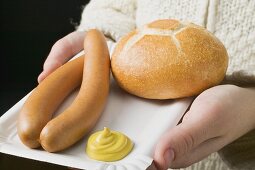 Person holding frankfurters, roll & mustard on paper plate