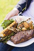 Woman holding plate of steak, grilled vegetables & corn on the cob