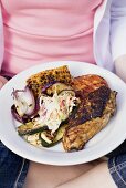 Seated woman holding plate of grilled chicken breast & accompaniments