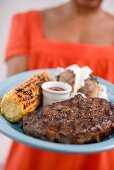 Woman holding plate of steak, corn on the cob, baked potato