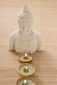 Three windlights in front of Buddha statue on bamboo mat