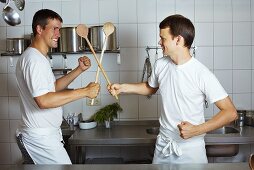 Two chefs fencing with wooden spoons in commercial kitchen