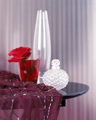 Rote Rose im rotem Glas, daneben ein Flakon und ein Seidentuch.
