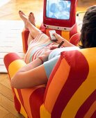 Frau auf Sessel vor TV, Fernbe- dienung, roter Fernseher