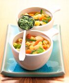 Two white bowls of minestrone and a spoon on blue tray