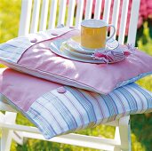 Geschirr in gelb auf Kissen in rosa und hellblau im Garten auf Stuhl