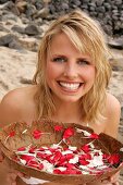 Pretty blonde woman sitting and holding bowl of white and red petals on beach, smiling