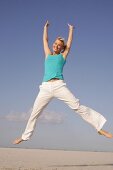 Pretty woman in blue top and white pants jumping on beach with arms outstretched, smiling