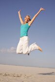 Pretty woman in blue top and white pants jumping on beach with arms outstretched, smiling