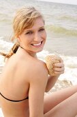 Pretty woman in black bikini rubbing her arm with sponge while sitting on beach, smiling