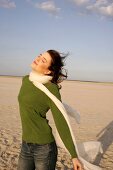 Carefree woman in green sweater, jeans and scarf enjoying sun on beach