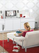Woman sitting on white sofa with sideboard watching TV
