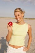 Woman holding a red apple standing on beach, laughing