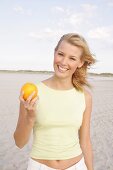 Frau hält in einer Hand eine Orange, sie steht am Strand