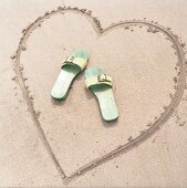In Sand gemaltes Herz mit zwei Sandaletten in der Mitte