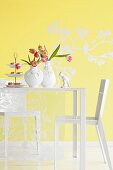Two vases of tulips and cake stand of confectionary on white table in front of yellow wall