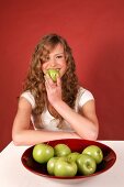 Happy woman sitting at table with bowl of green apples and holding apple in hand