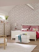 White double bed and chests of drawers in bedroom with pink and white floral wallpaper and ceiling beam