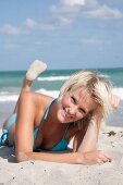 Portrait of beautiful blonde woman wearing blue bikini lying on beach, smiling