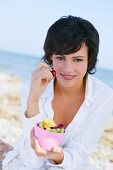 Brunette woman wearing white shirt sitting on beach while having fruit from bowl