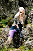 blonde Frau sitzt auf Steinen, lacht lila Strumpfhose, Pelzweste.