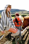 Two beautiful women wearing knitted poncho, hood and cap standing near the fence