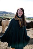 Happy brunette woman wearing poncho and scarf standing in pasture and laughing