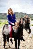 Pretty blonde woman in blue sweater riding a black icelandic horse, looking over shoulder