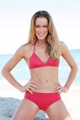 Portrait of pretty blonde woman in red bikini posing with hands on hip at beach, smiling