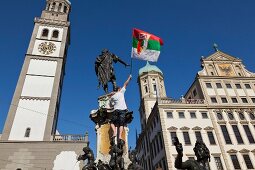 Man holding flag standing on Augustus fountain in front of Perlachturm, Augsburg, Germany