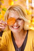 Happy blonde woman in yellow sweater holding a autumn leaf in front of left eye, smiling