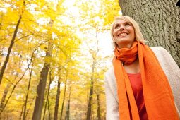 Blonde woman wearing scarf and cardigan leaning against tree in autumn forest, smiling