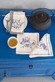 A teapot, a tea bowl and linen napkins embroidered with various motifs on a blue chest of drawers